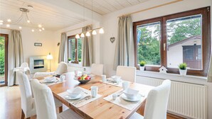 A dining area with a wooden table set for four, white chairs, and a view of a house through large windows.