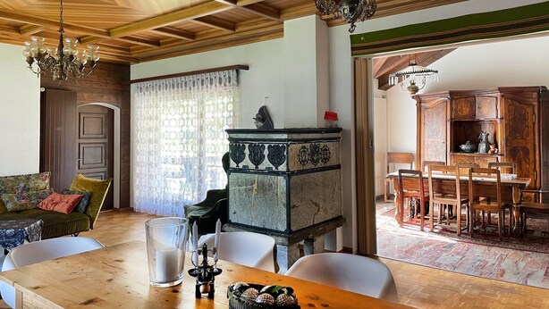 A dining area with a wooden table, chairs, and a chandelier. There is a wooden cabinet with decorative elements and a window with patterned curtains. - Fifinhaus 1. OG Süd by Interhome (Fiesch)