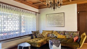 A cozy living room with a wooden ceiling, a large window with patterned curtains, a sofa with multiple cushions, a small side table, and a chandelier. - Fifinhaus 1. OG Süd by Interhome (Fiesch)