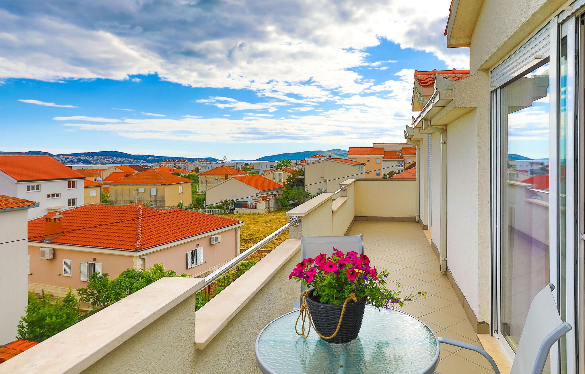 Un balcón con una mesa de vidrio y una planta en maceta, con vistas a una zona residencial de casas con tejados rojos.