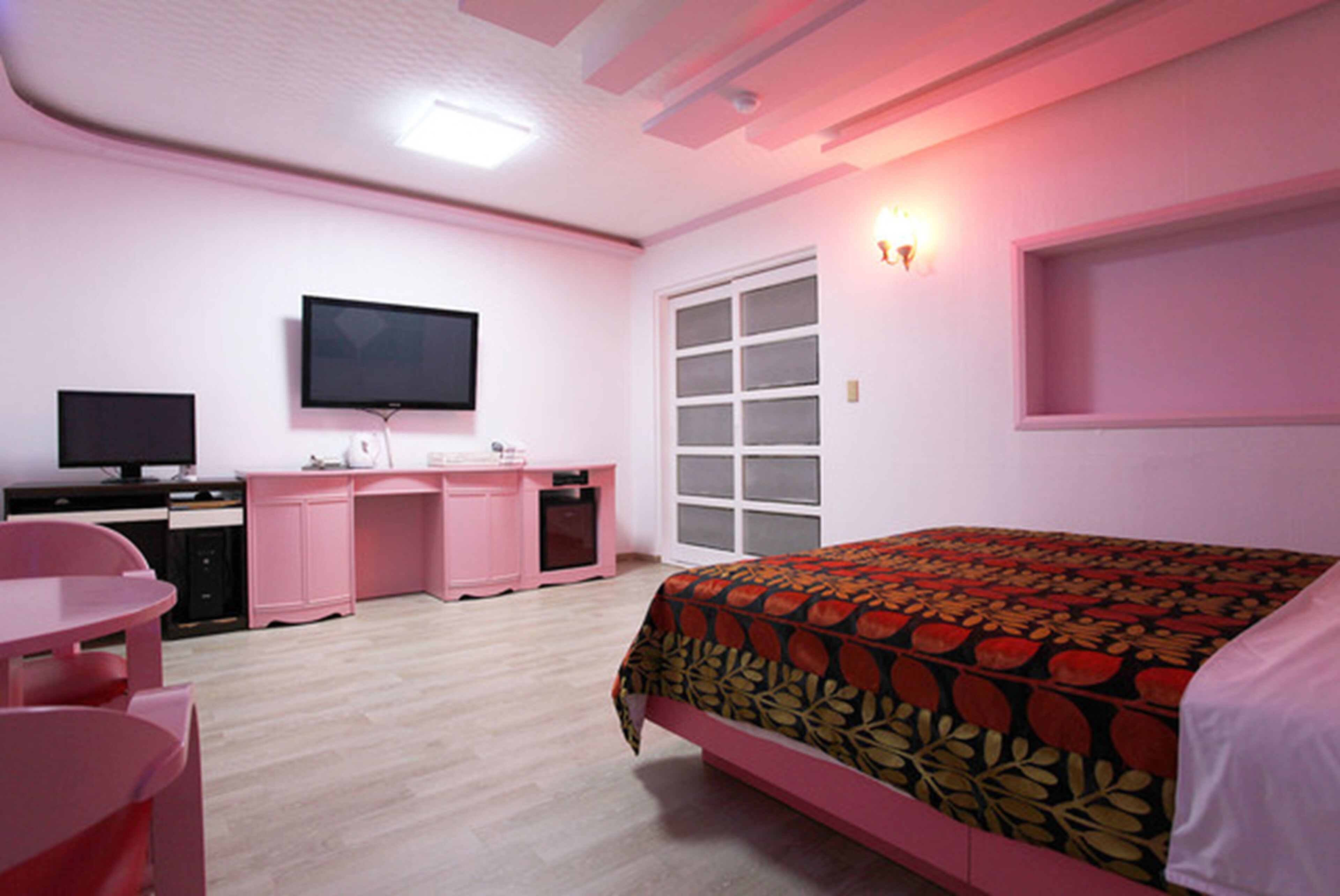 Un dormitorio con decoración rosa que incluye una cama, un escritorio, una silla, un televisor y una ventana.