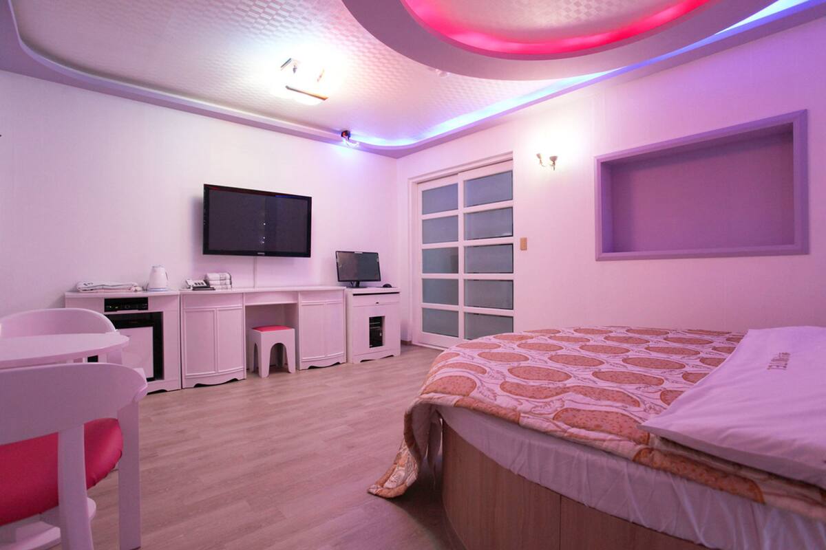 A bedroom with a bed, a desk, a television, and a window.
