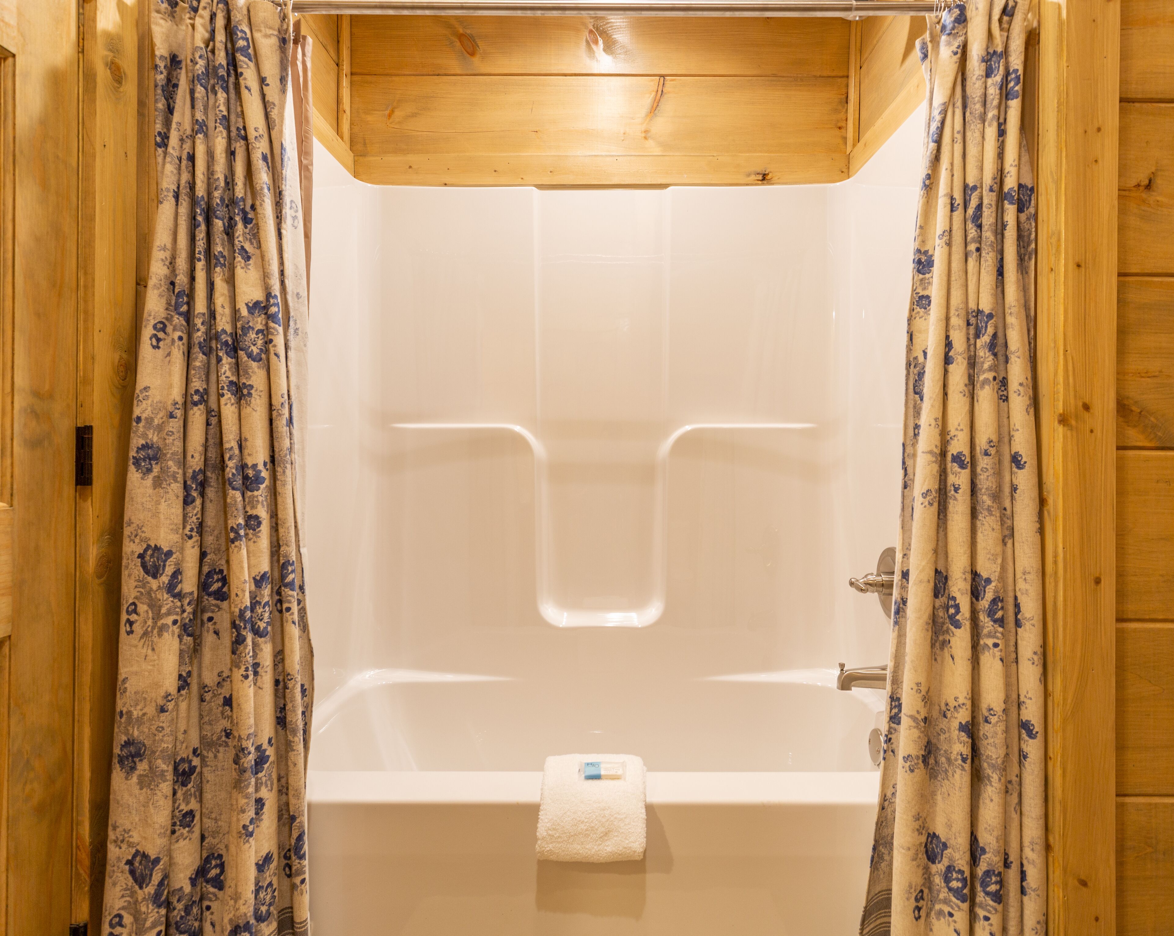 Una ducha con estructura de madera, una bañera blanca, cortinas con motivos florales y una toalla colgada de la bañera.