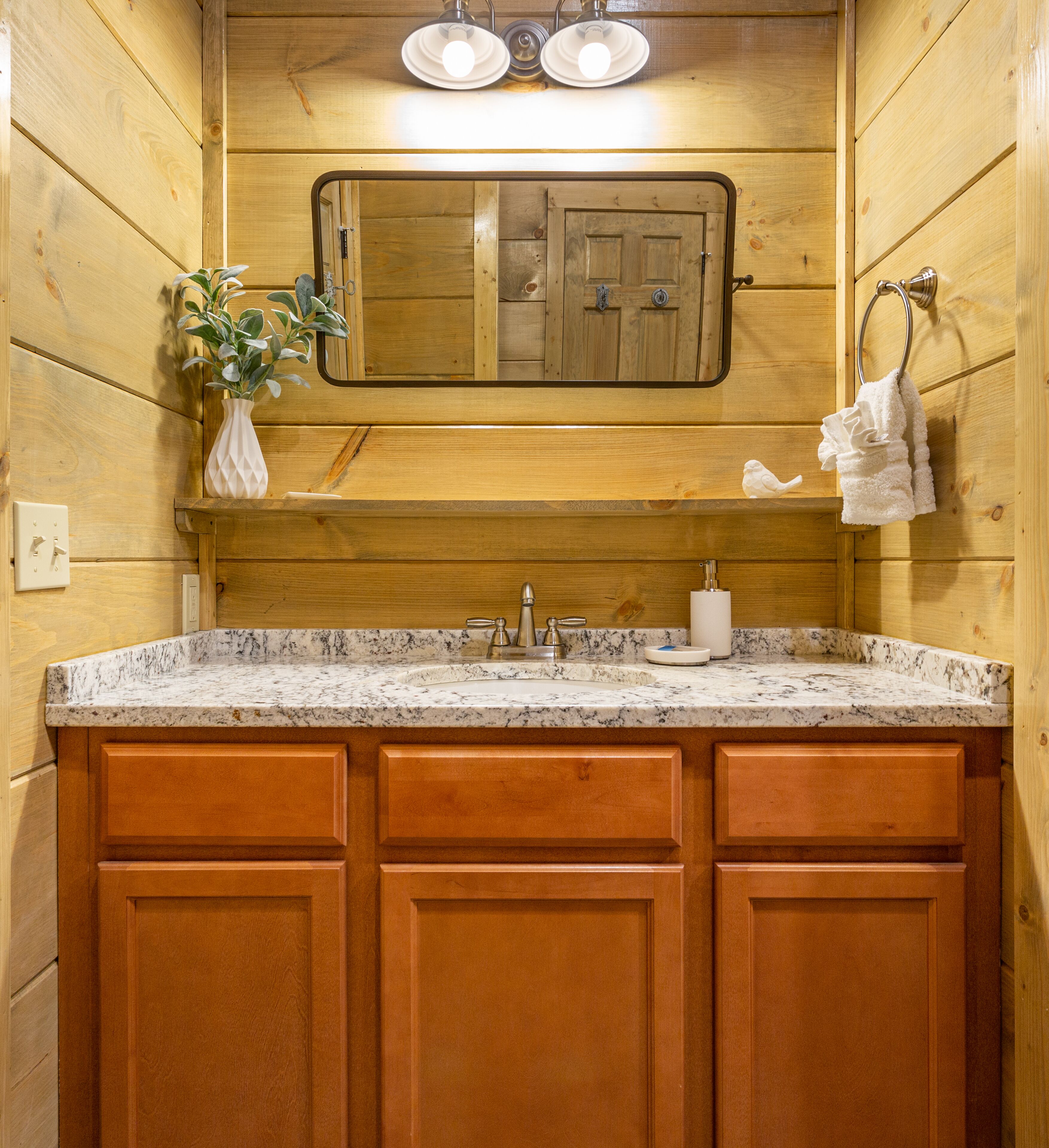 Un baño estilo cabaña de madera con lavamanos de granito, armarios de madera, un espejo y un perchero para toallas.