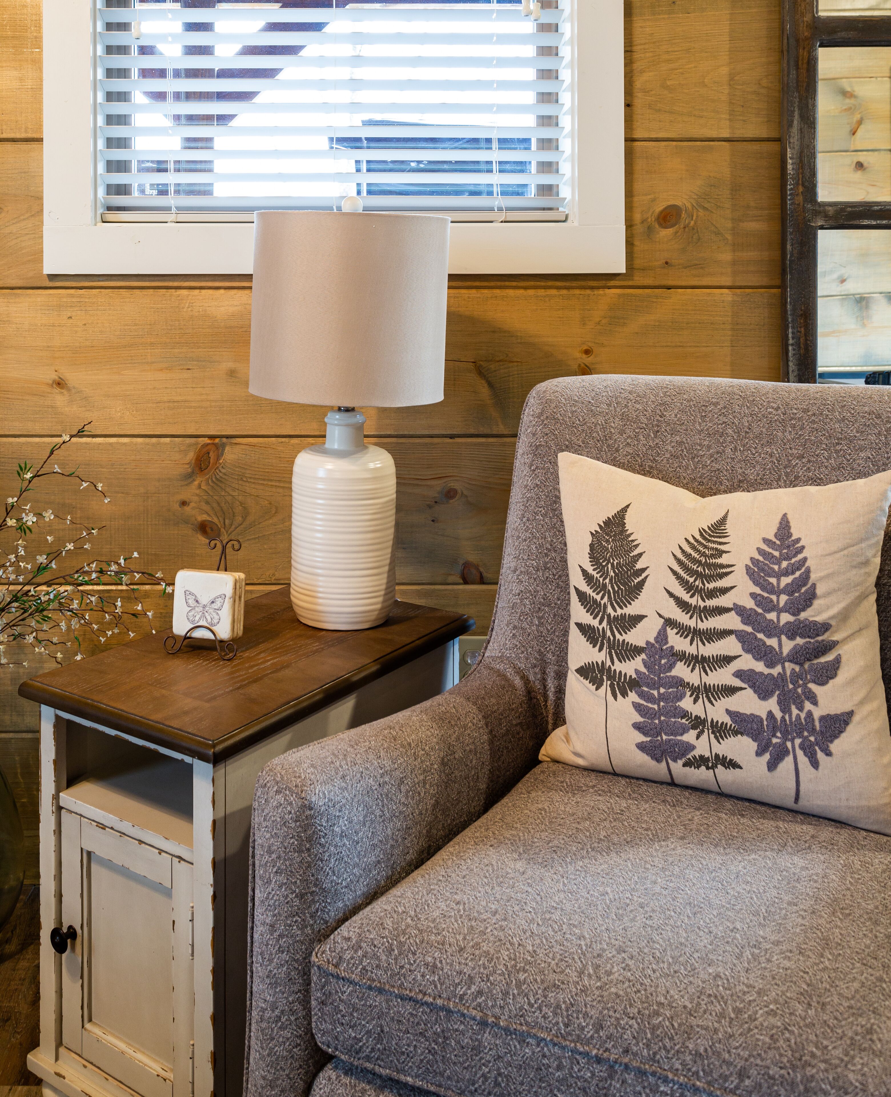 A cozy room with a wooden wall, a window with blinds, a lamp on a wooden side table, a gray sofa, and a decorative pillow with fern designs.