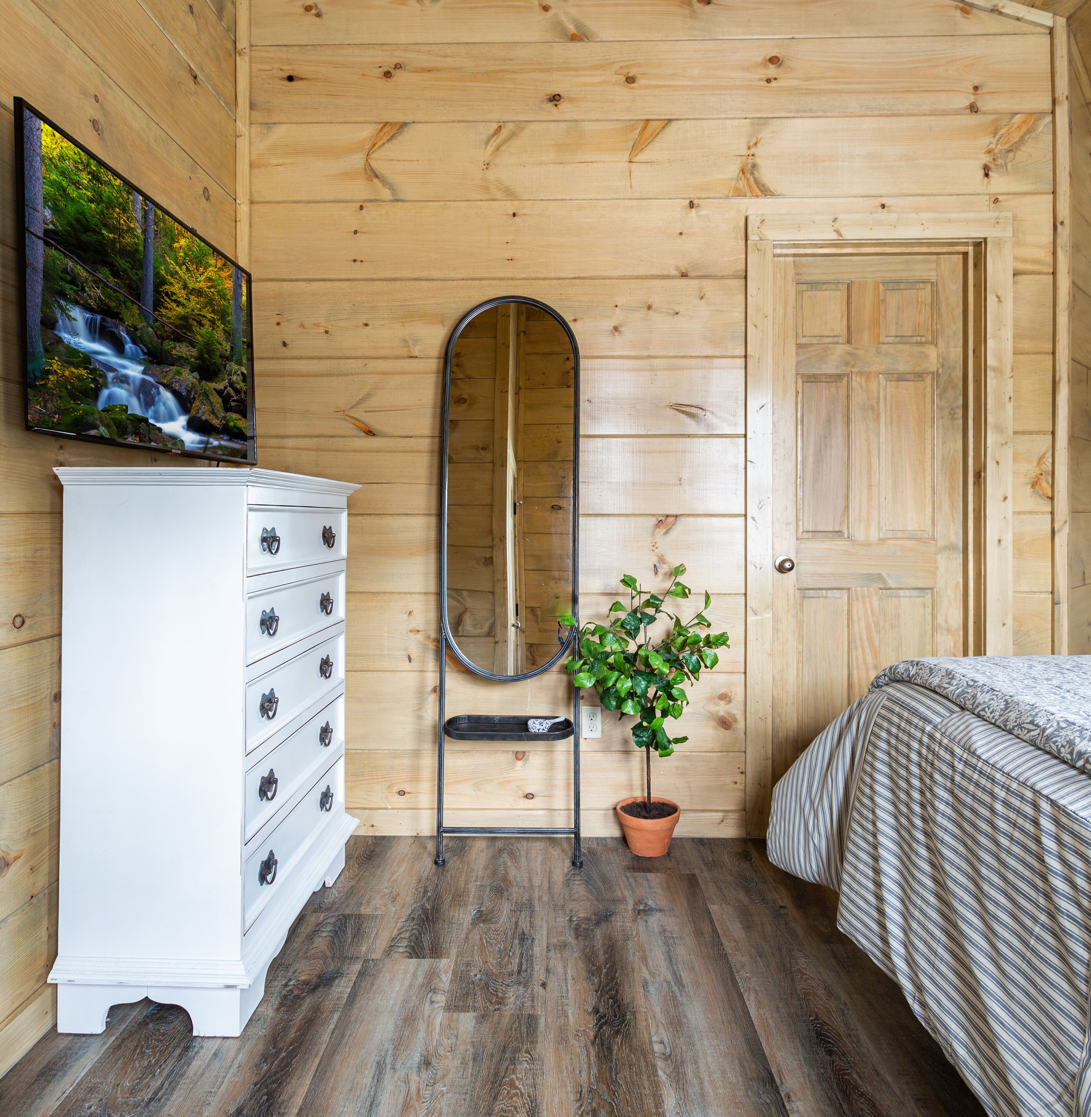 Ein Zimmer aus Holz mit einem Bett, einem Schminktisch, einem Spiegel und einer Zimmerpflanze.