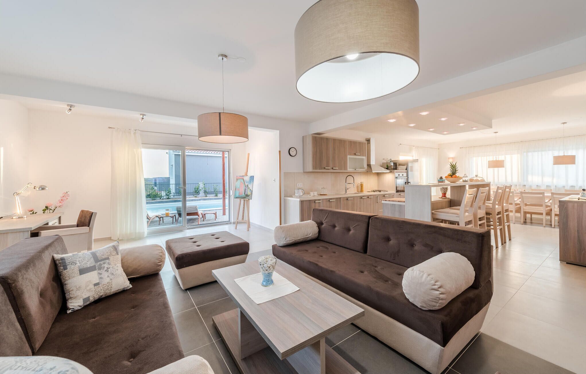 A modern living room with a sofa, ottoman, and coffee table, a dining area with chairs, and a kitchen in the background.