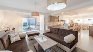 Un salon moderne avec un canapé, un pouf et une table basse, un coin repas avec des chaises, et une cuisine en arrière-plan.
