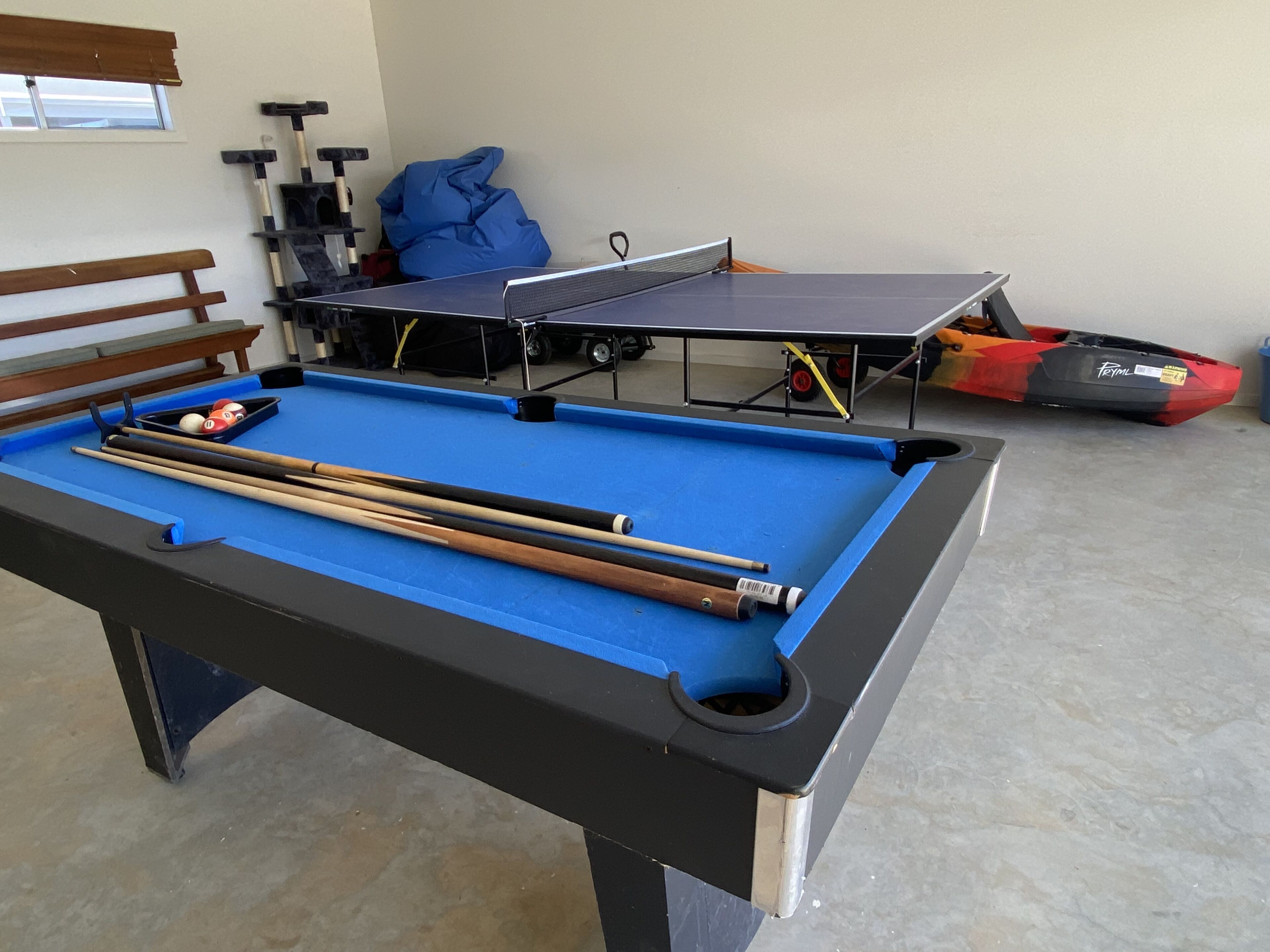 Ein Billardtisch mit Billardkugeln und Spielstangen, ein Tischtennis-Tisch und ein Kajak in einem Raum mit einer Bank und einem Fenster.
