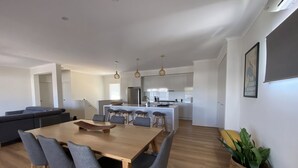 A modern kitchen with white cabinets, a central island with stools, and a dining area with a wooden table and grey chairs. - Room to relax in a perfect location with an easy walk to the beach and shops. (Normanville)