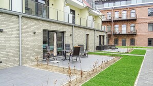 An outdoor seating area with tables and chairs, a small garden with grass and plants, and a brick building in the background. - Cozy apartment in Malchow with WiFi (Malchow)