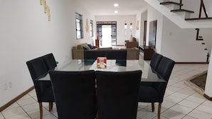 A dining area with a glass table surrounded by four chairs, a sofa in the background, and a staircase leading upstairs. - Your best season in Gabriela's land (Ilhéus)