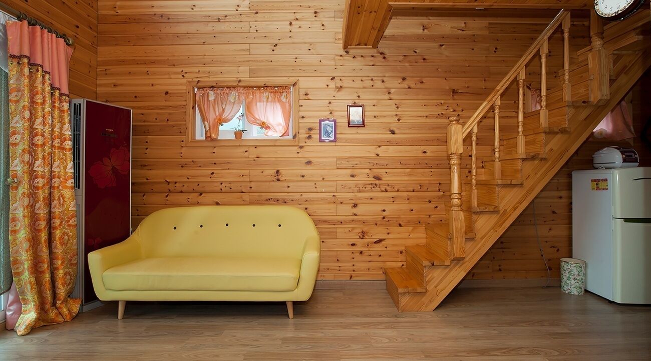 A wooden cabin interior with a yellow sofa, a staircase, a window with curtains, and a refrigerator.
