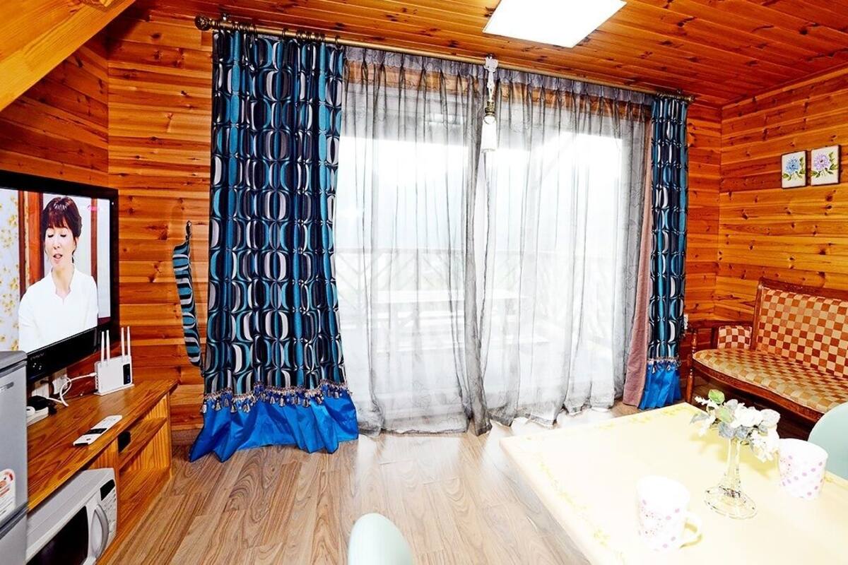 A cozy cabin-style room with a wooden interior, a checkered sofa, a dining table with a vase of flowers, and a television on a wooden stand.