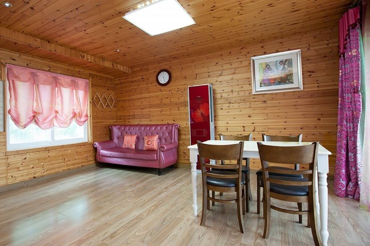 A wooden cabin interior with a dining table, chairs, a pink sofa, a clock, and a window with curtains.