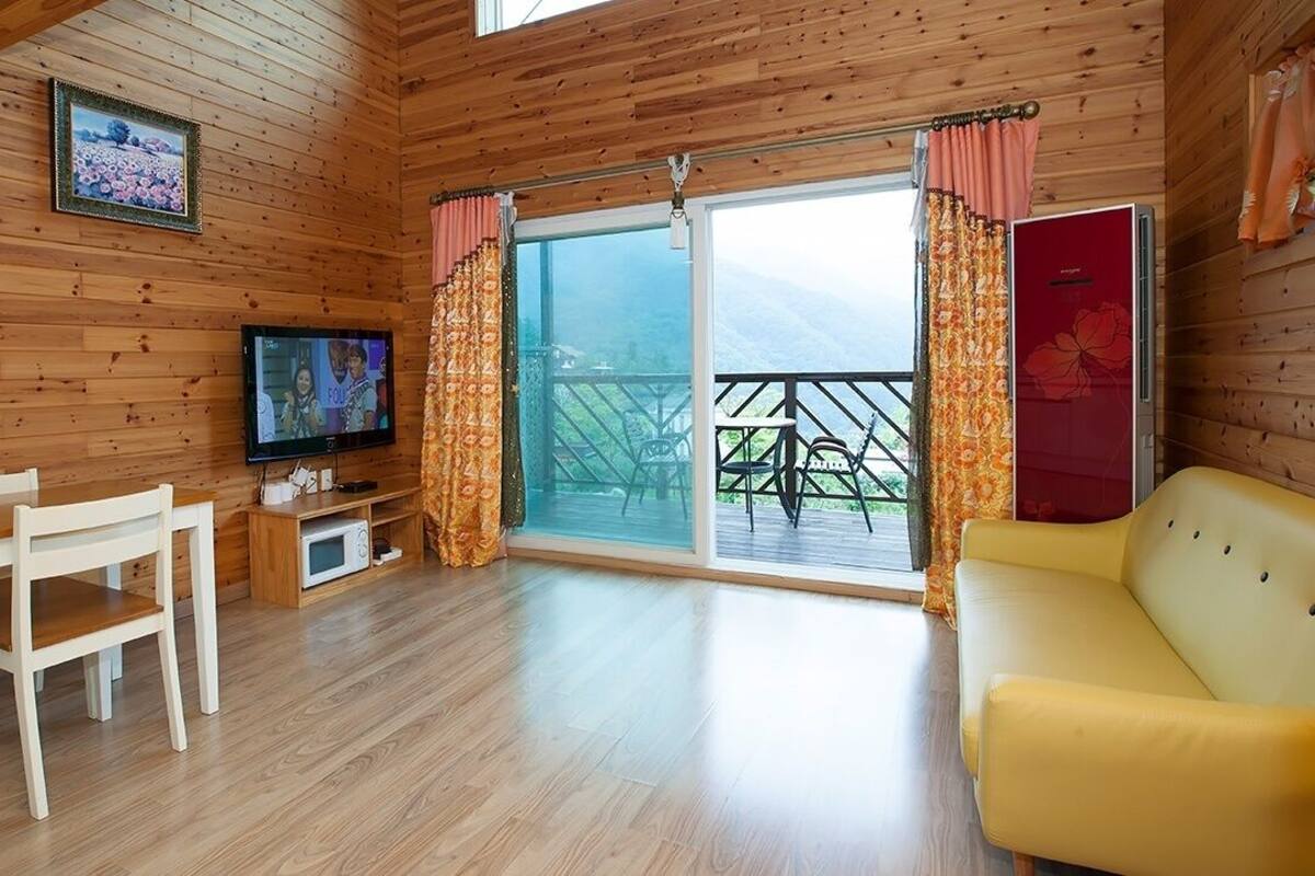 A wooden cabin with a TV, a yellow sofa, a dining table, and a balcony with a view of mountains.