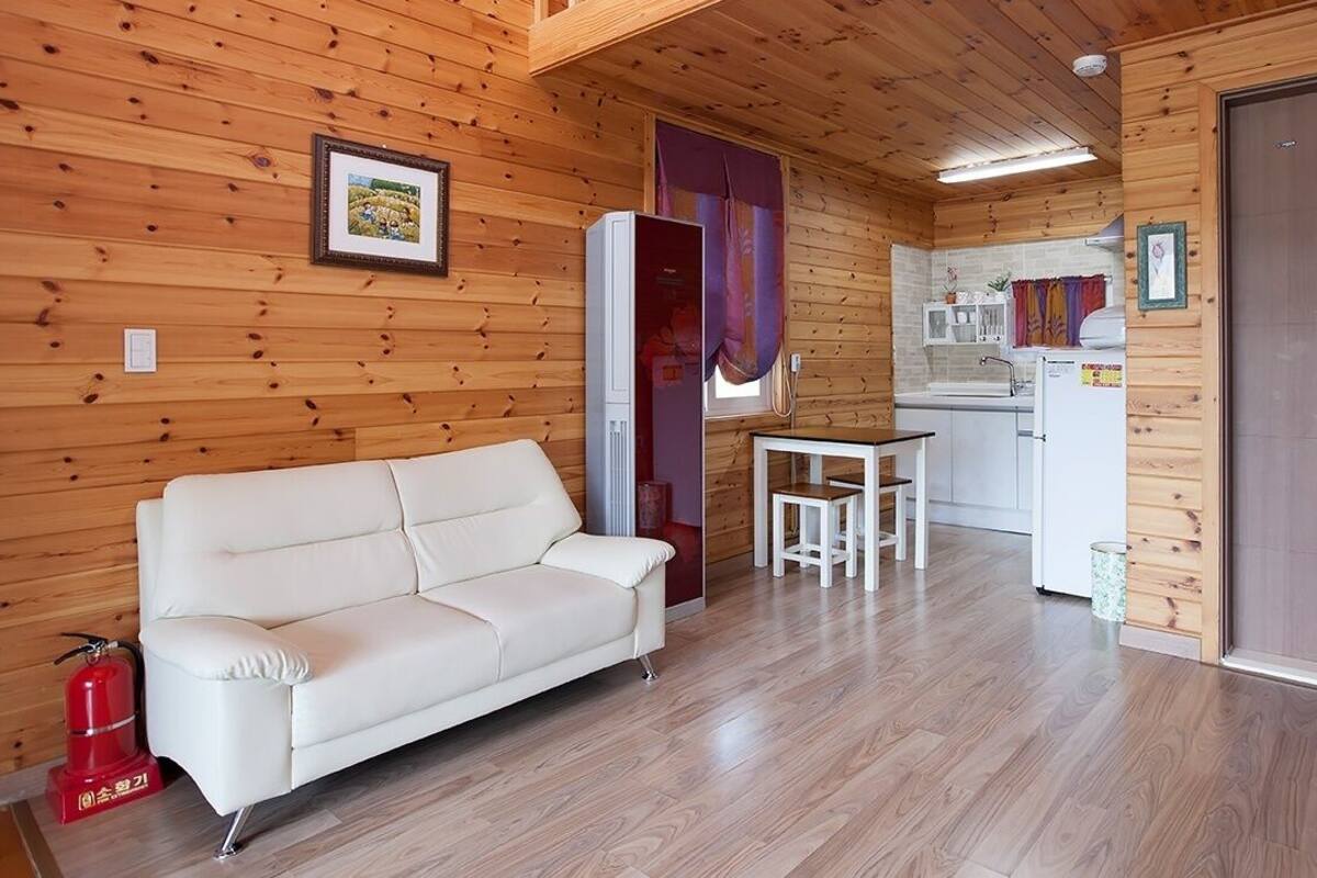 A wooden-paneled room with a white sofa, a small table, and a kitchen area with a refrigerator and sink.