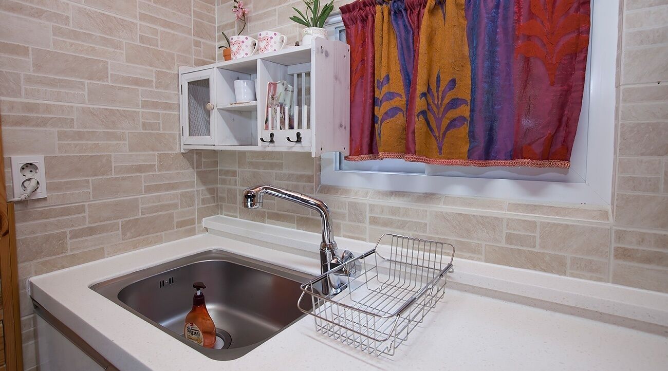 A kitchen with a stainless steel sink, a white countertop, a metal drying rack, a window with a colorful curtain, and a shelf with decorative items.
