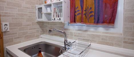 A kitchen with a stainless steel sink, a white countertop, a metal drying rack, a window with a colorful curtain, and a shelf with decorative items.
