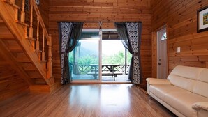A wooden cabin interior with a wooden staircase, a beige sofa, and a large sliding glass door leading to a balcony with a picnic table. - Laviche Pension (Gapyeong)