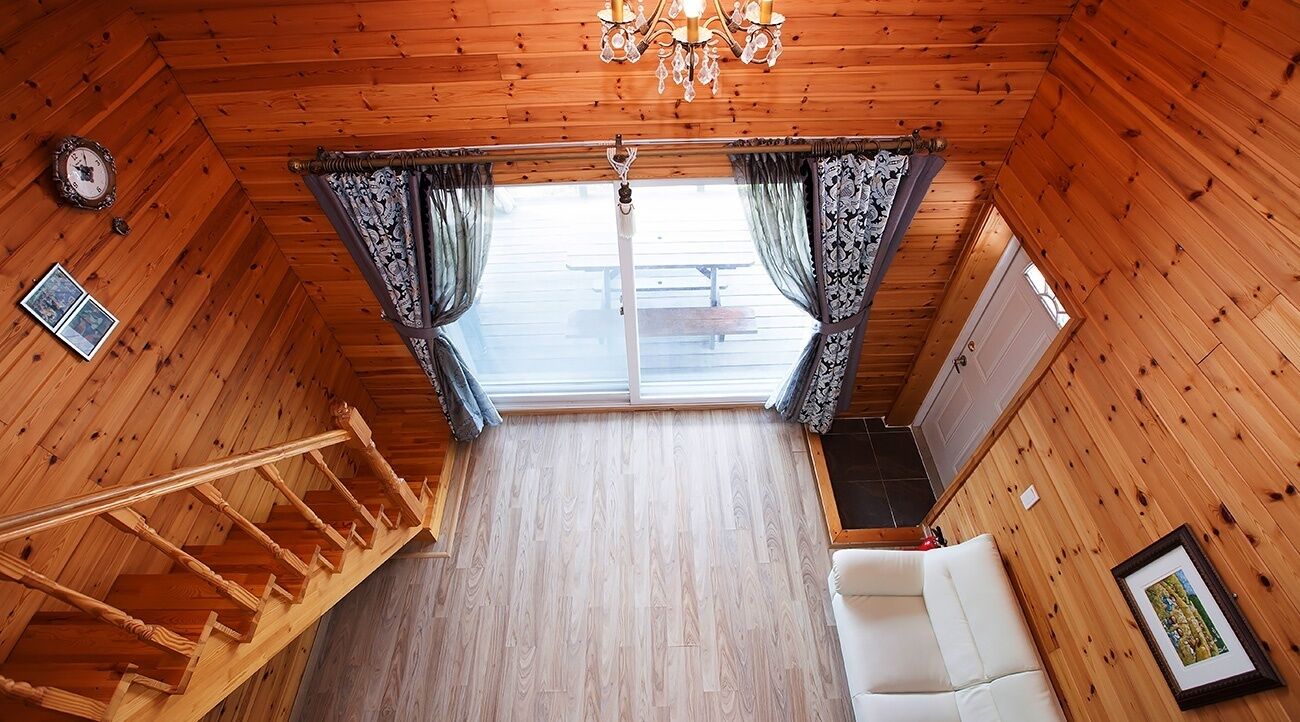 A wooden cabin interior with a staircase, white sofa, chandelier, and a view through a window.