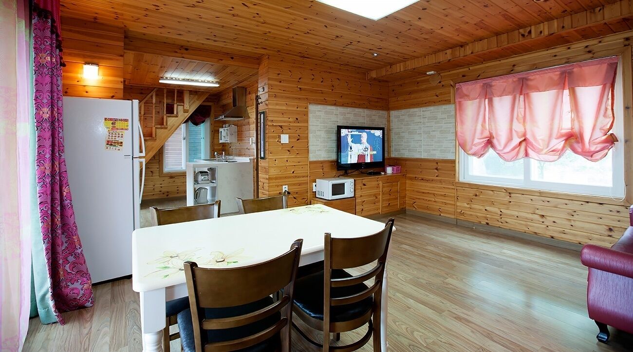A wooden cabin interior with a dining table, chairs, a TV, a microwave, and a staircase leading to a kitchen.