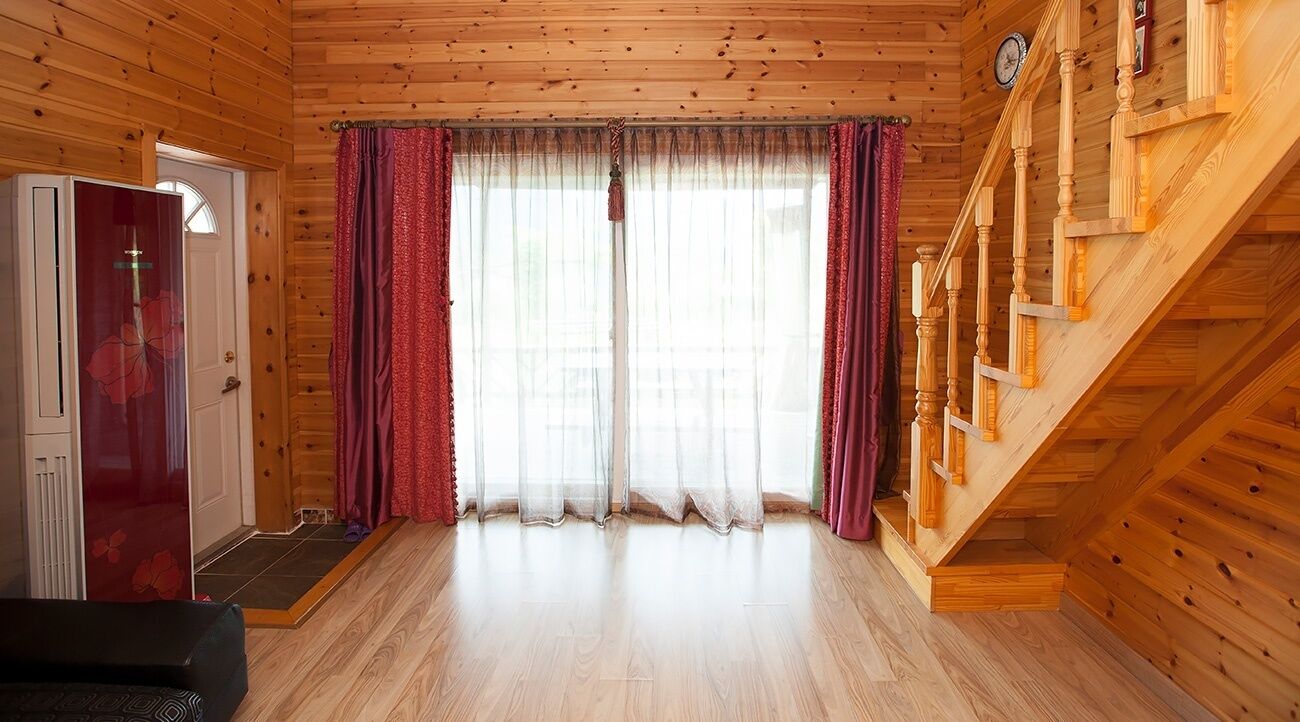Interior de una cabaña de madera con una escalera, una puerta con panel de vidrio y una ventana con cortinas translúcidas.