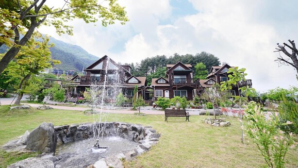 A large wooden house with multiple balconies, a fountain, and a bench in a landscaped garden. - Laviche Pension (Gapyeong)