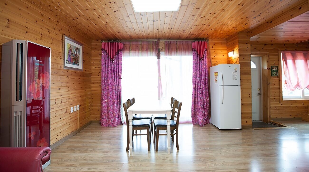 A wooden cabin with a dining area featuring a table and chairs, a refrigerator, and a large window with curtains.