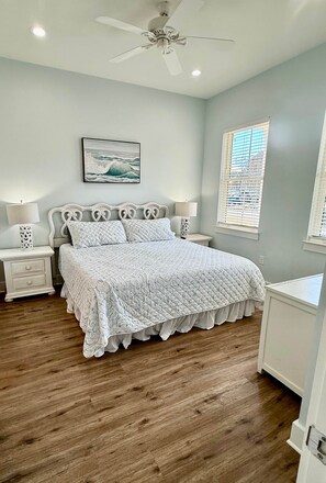 A bedroom with a bed, two bedside tables, a ceiling fan, a window with blinds, and a wall-mounted picture.