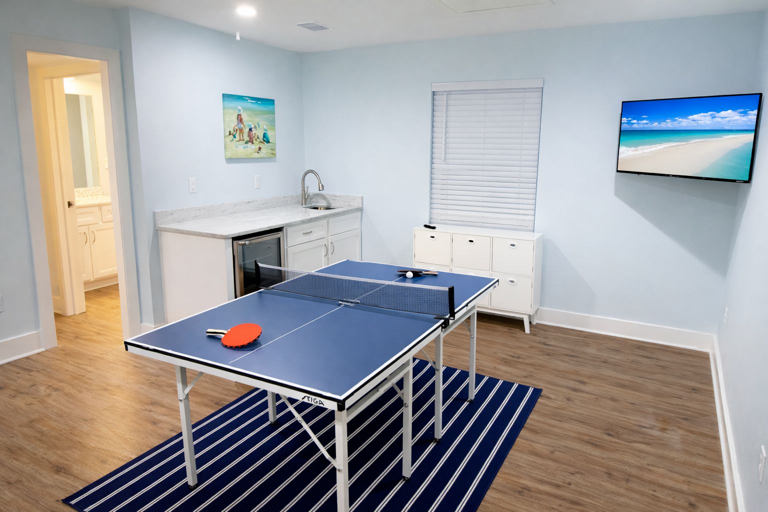 A room with a ping pong table, a kitchen area, a sink, a window with blinds, and a TV on the wall.