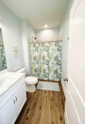 Un bagno con il water, un mobile lavabo con uno specchio, una tenda della doccia con motivo a foglia e un pavimento in legno.