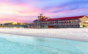 Un edificio fronte spiaggia con tetto rosso, diversi balconi e un pontile di legno che si estende sull'acqua.