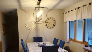 Uma área de jantar com uma mesa de madeira, cadeiras azuis, toalha de mesa florida, um relógio de parede e uma janela com cortina.
