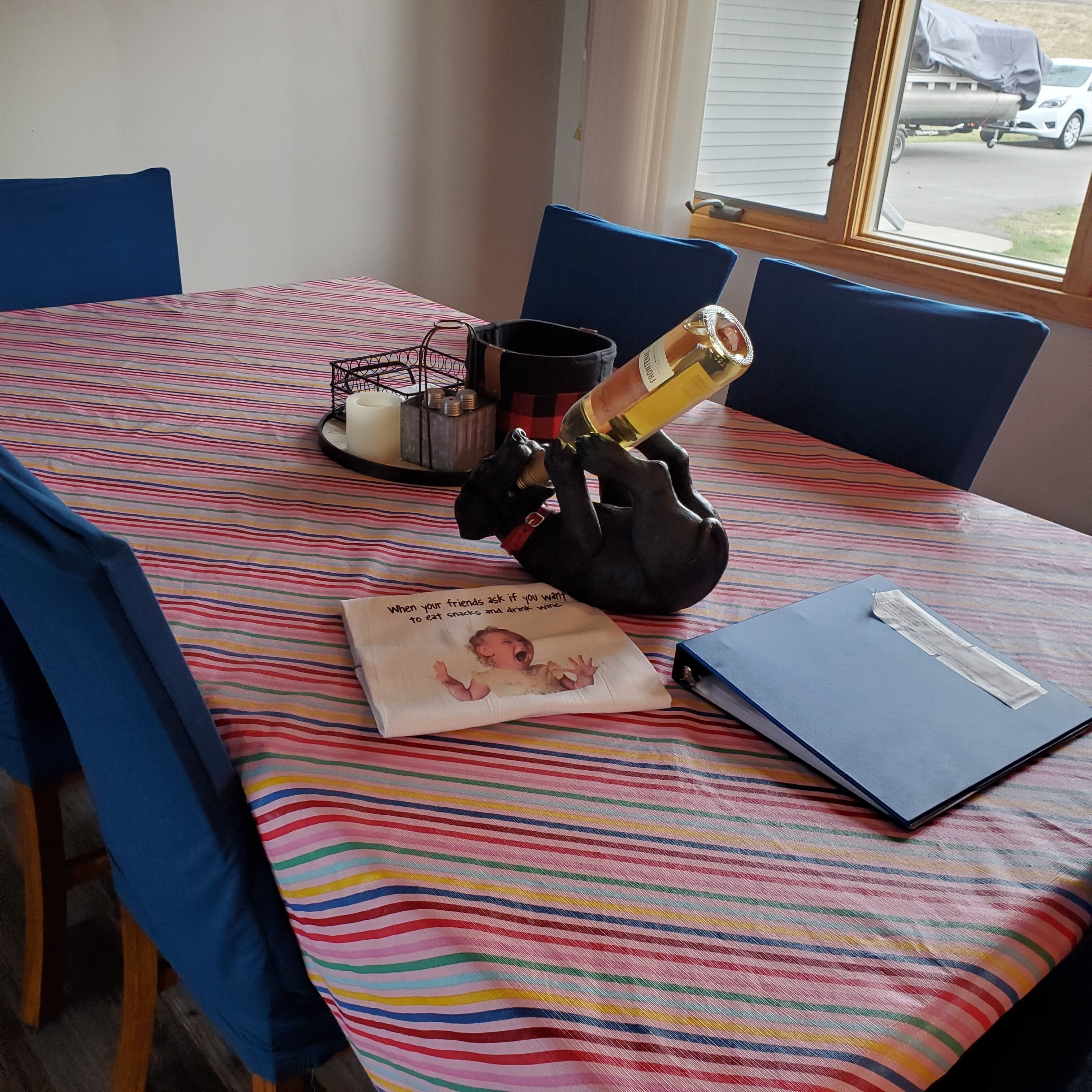 A table with a striped tablecloth, a candle, a metal candle holder, a bottle, a black sculpture, a book, and a window with a view of a vehicle outside.