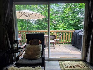 Een kamer met een schuifglasdeur die uitkomt op een terras met houten reling, een comfortabele stoel en een tafel.