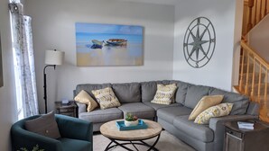 Uma sala de estar com um sofá modular cinza, uma mesa de centro redonda, uma poltrona azul-esverdeada, uma mesinha de apoio e um quadro com barcos na parede.