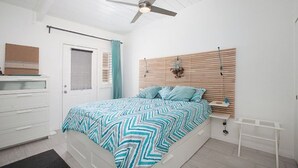 Quarto com cama, ventoinha no teto, cabeceira de madeira, cómoda branca e janela com cortinas.
