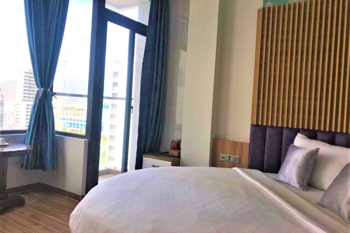 A hotel room with a large bed, a wooden headboard, a nightstand, a desk with a chair, and a window with curtains.
