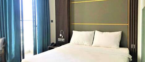 Quarto de hotel com uma cama grande, uma mesinha de cabeceira com telefone e vista para o exterior através de uma janela com cortinas azuis.