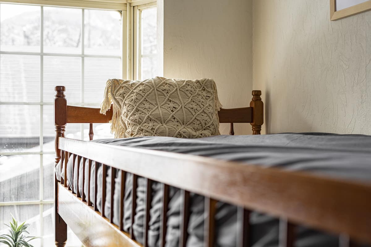 A wooden bed with a patterned throw blanket, a window with blinds, and a view of a snowy landscape.