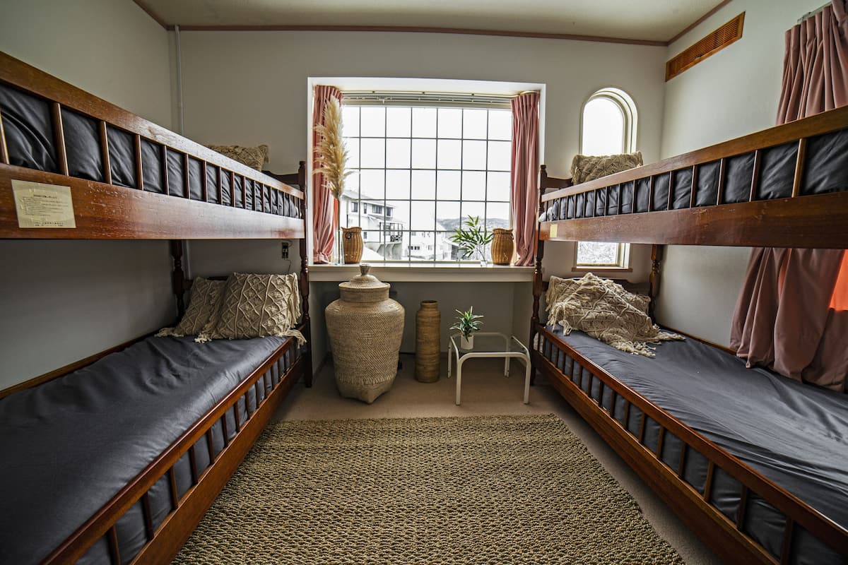 A room with bunk beds, a window with curtains, a small table, and a rug.