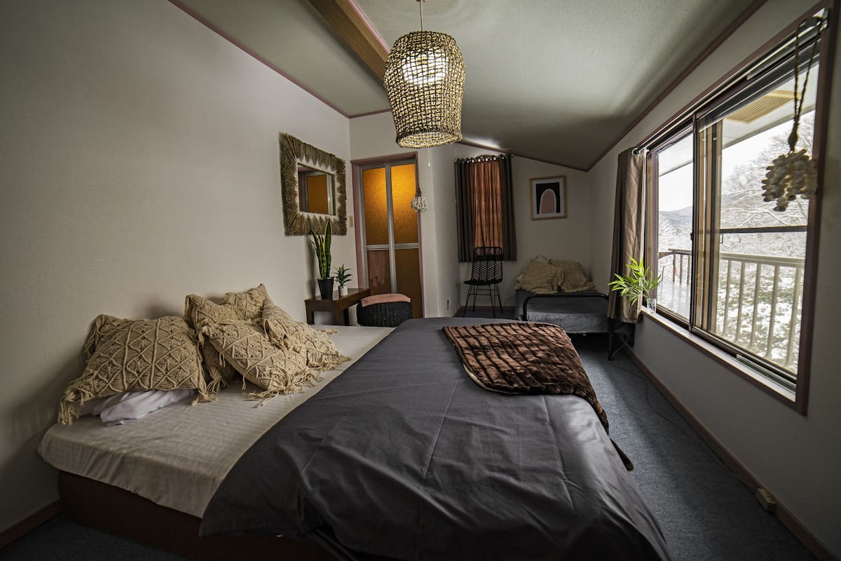 A bedroom with a bed, a bedside table, a chair, a window with a view, and a hanging light fixture.