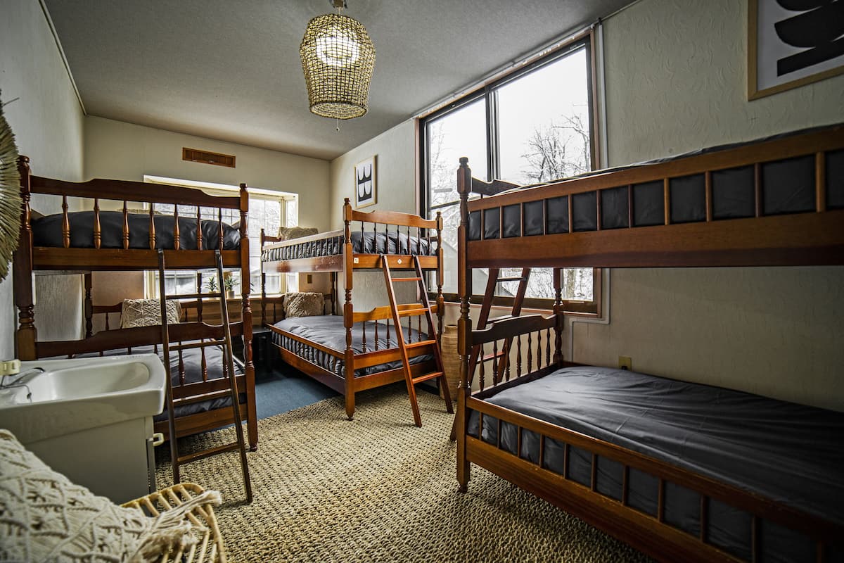 A dormitory room with bunk beds, a ladder, a sink, and a window with a view of trees.
