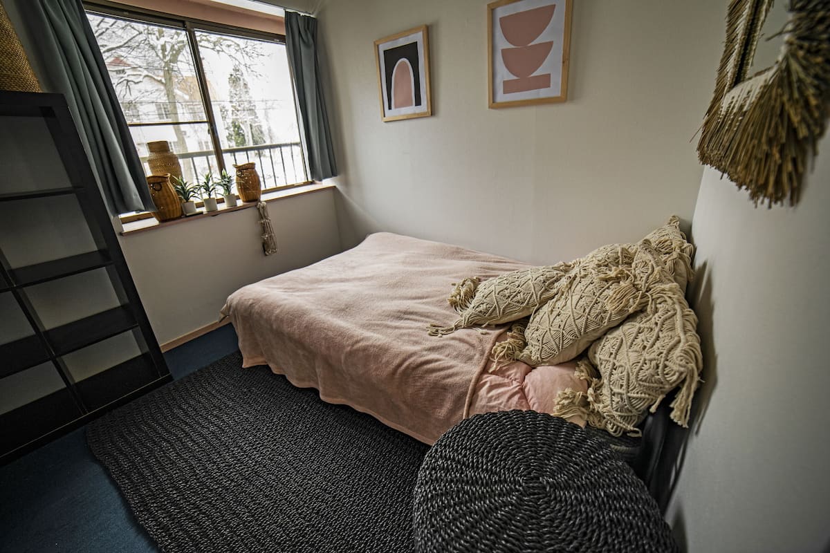 A bedroom with a bed, a window with curtains, a small table with decorative items, and a wall-mounted shelf.