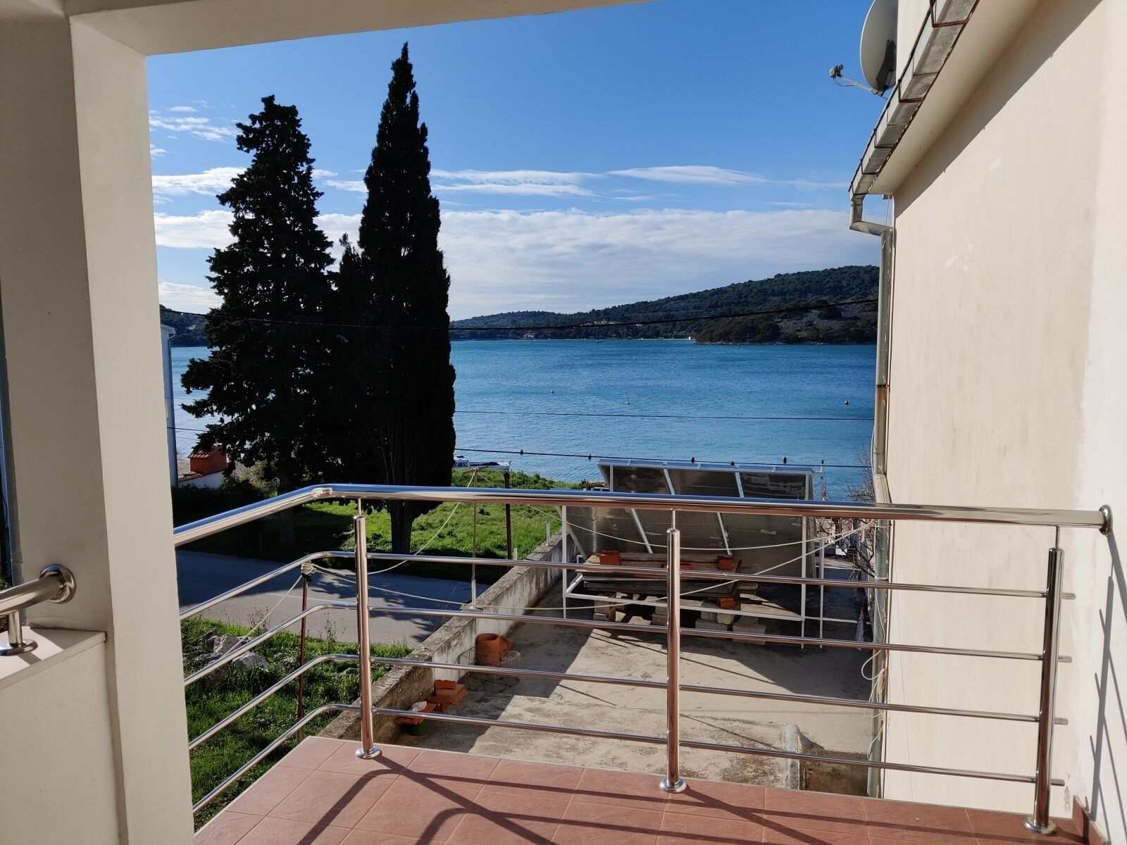 A balcony with a metal railing overlooking a body of water, a tree, and a distant hill.