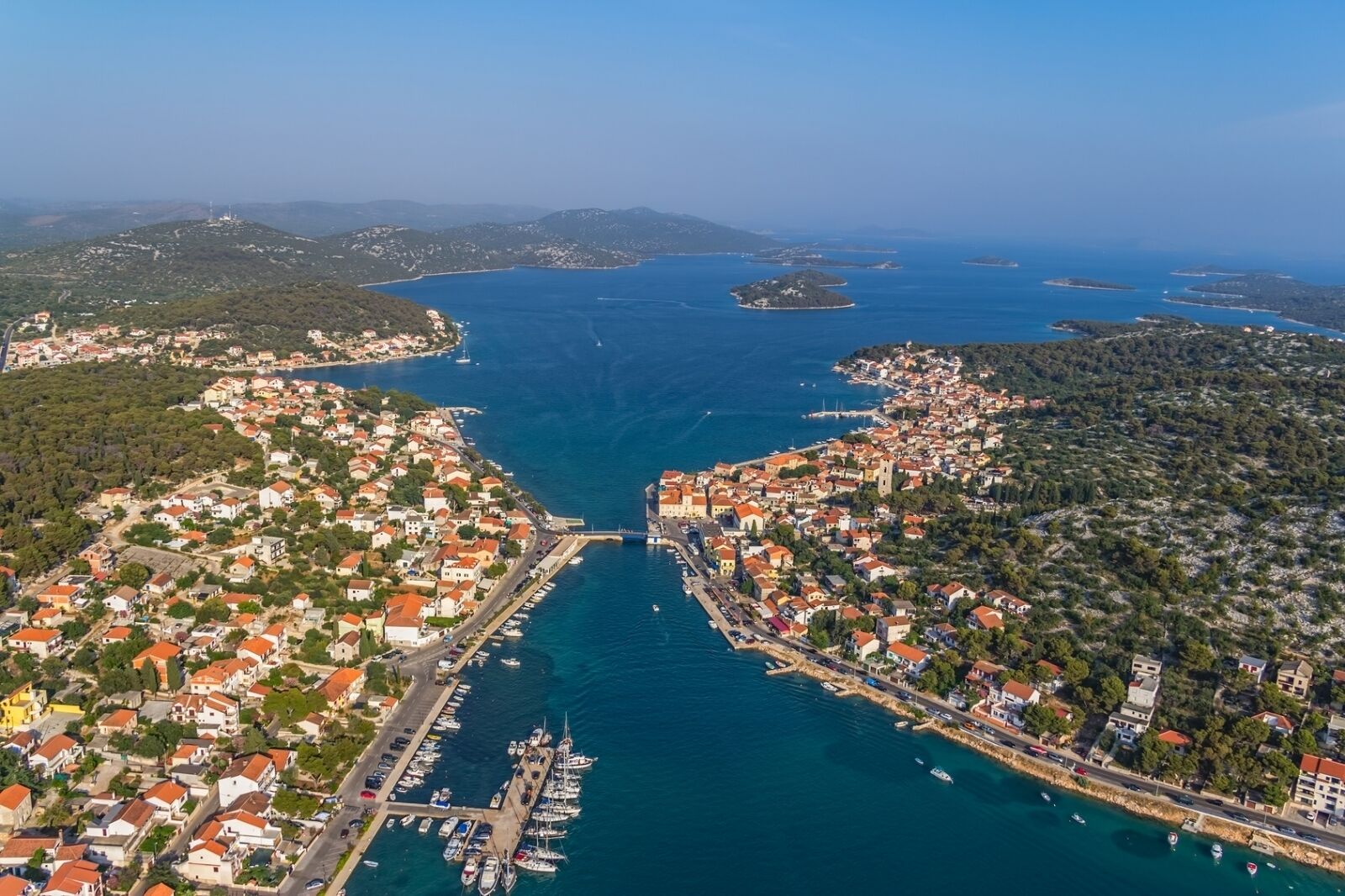 Vista aerea di una località costiera con un porto turistico, edifici fitti e un mare cristallino.