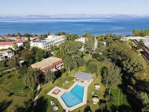 Ein Luftbild zeigt einen Urlaubsort mit einem Schwimmbad, das von Grünflächen umgeben ist, ein großes mehrstöckiges Gebäude und den Blick auf das Meer.