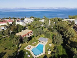 An aerial view of a resort with a swimming pool surrounded by greenery, a large building with multiple floors, and a view of the sea.