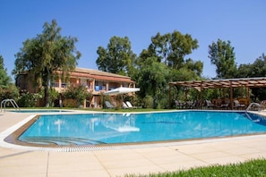 A swimming pool with lounge chairs and a covered area, surrounded by trees and a building with a red roof. - Tellis Holiday Rooms (Corfu)